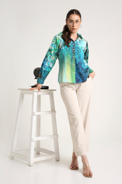 A woman wearing a long-sleeve button-up shirt with a vibrant blue, green, and teal abstract print, paired with cream-colored trousers, standing next to a white wooden stool against a plain white studio background.