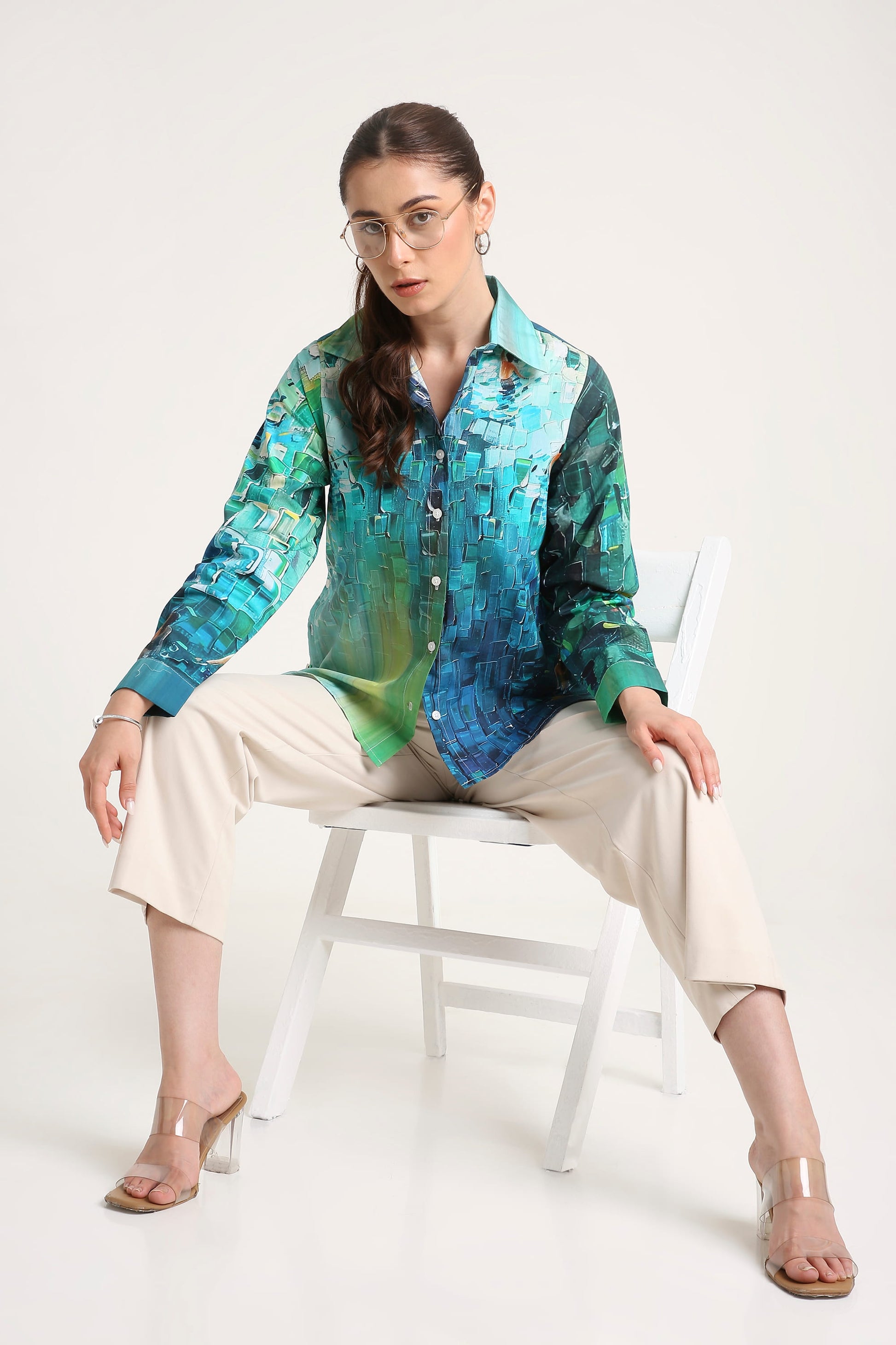 A woman wearing a long-sleeve button-up shirt with a vibrant blue, green, and teal abstract pattern, paired with beige cropped pants and clear high-heeled sandals, sitting on a white chair against a light gray background.
