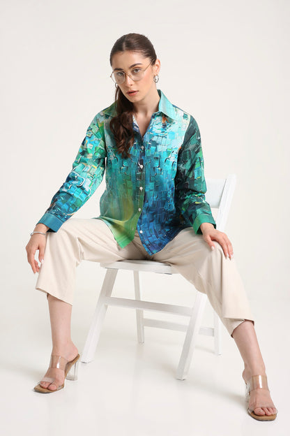 A woman wearing a long-sleeve button-up shirt with a vibrant blue, green, and teal abstract pattern, paired with beige cropped pants and clear high-heeled sandals, sitting on a white chair against a light gray background.