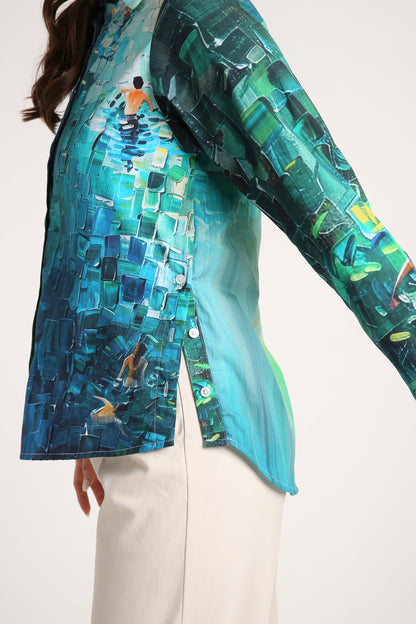 A close-up of a woman wearing a long-sleeve button-up shirt with a vibrant blue and green mosaic print depicting figures in water, paired with off-white trousers.
