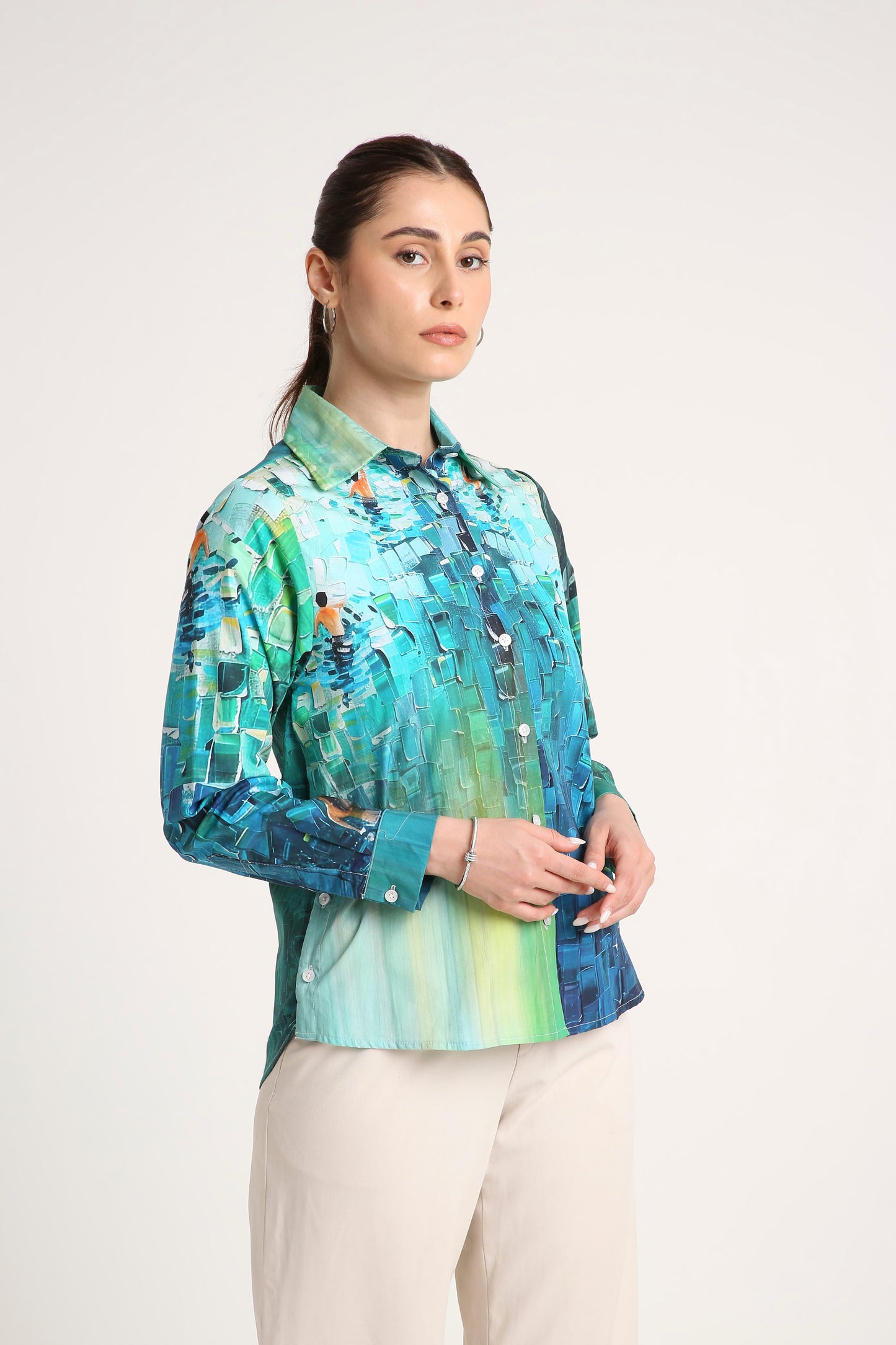 A woman wearing a long-sleeve button-up shirt with an abstract painterly pattern in shades of teal, blue, and green, paired with light beige trousers, against a plain off-white studio background.