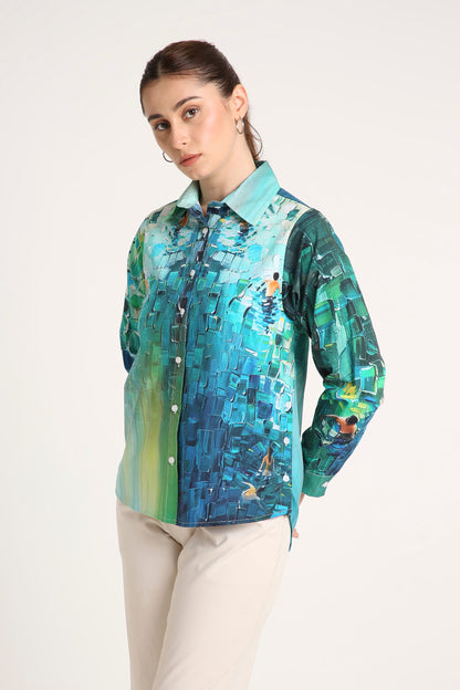 A woman wearing a long-sleeve button-up shirt with a vibrant blue, green, and yellow abstract mosaic print, paired with beige trousers, against a plain light background.