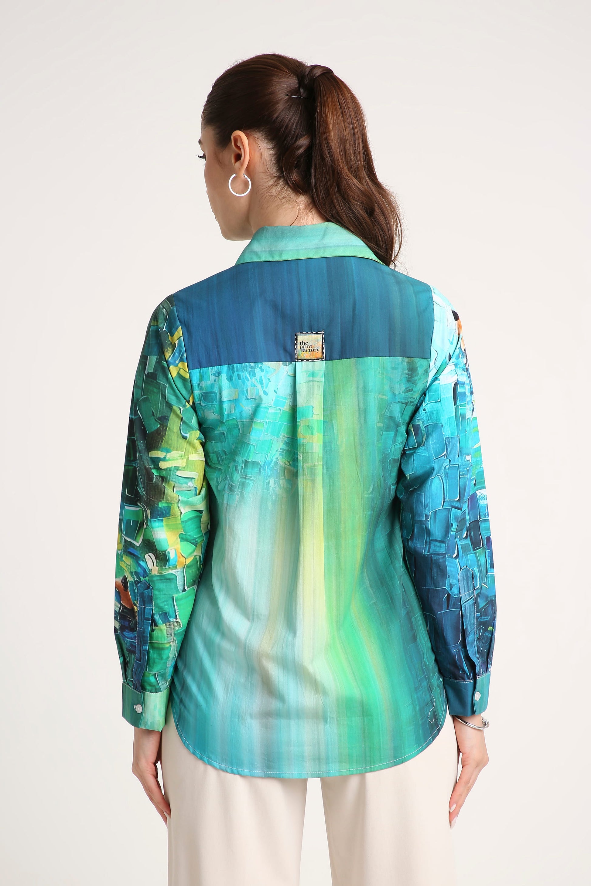 A woman viewed from the back wearing a long-sleeve collared button-up shirt with an abstract painterly print in shades of teal, blue, green, and yellow, paired with light beige trousers against a plain off-white background.