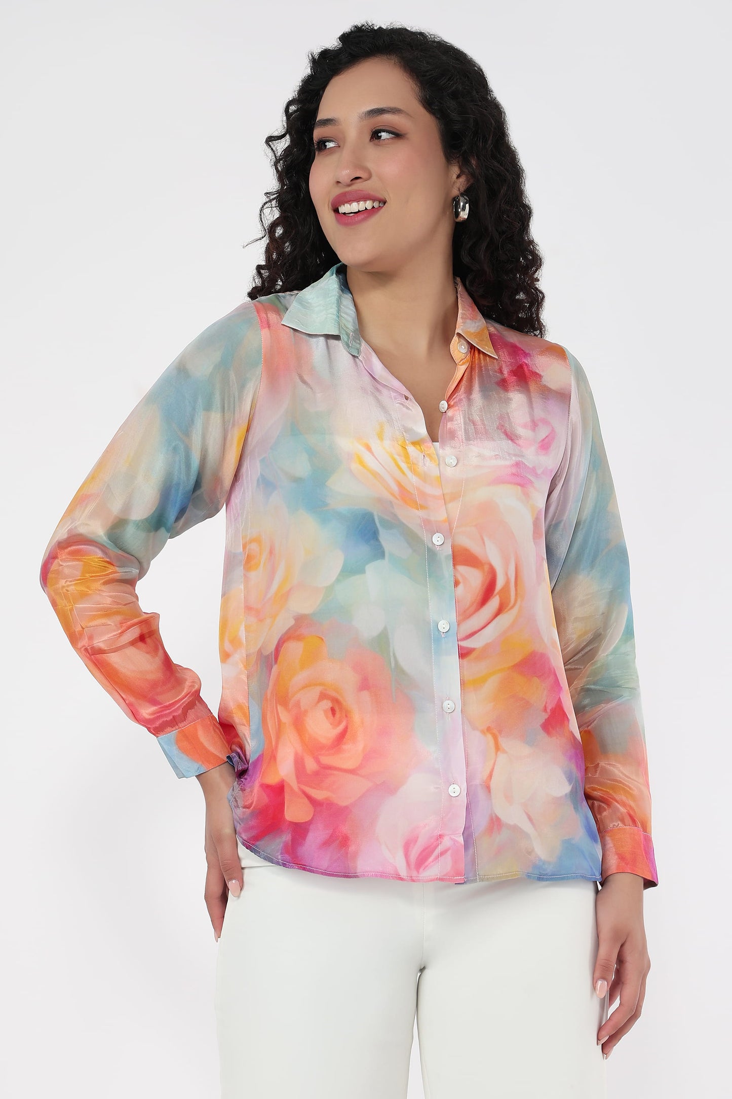 A woman wearing a long-sleeve button-up shirt with a colourful watercolour floral print, paired with white pants, against a plain light grey background.