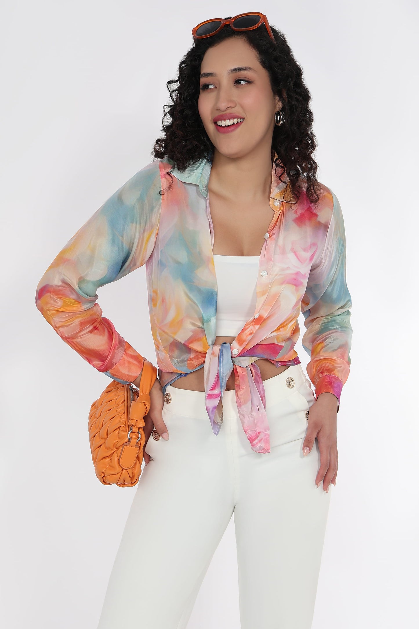 A woman modeling a lightweight, long-sleeve tie-front shirt with a vibrant pastel tie-dye pattern in blue, orange, pink, and yellow, paired with white pants and an orange textured clutch bag.