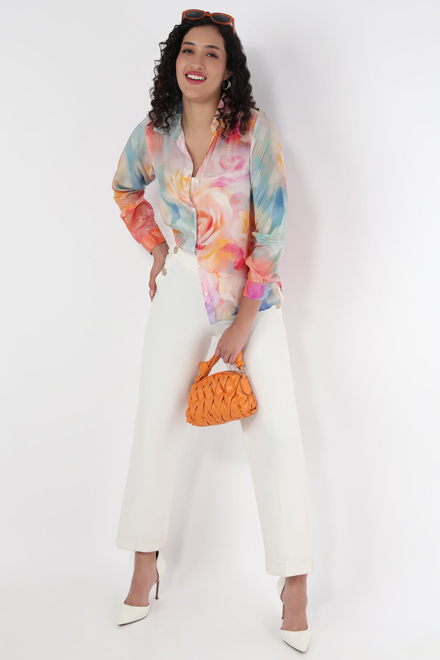 A woman modeling a long-sleeve button-up shirt with a vibrant pastel watercolor floral print, paired with white wide-leg cropped pants, white pointed-toe heels, and a small orange woven handbag against a plain white studio background.