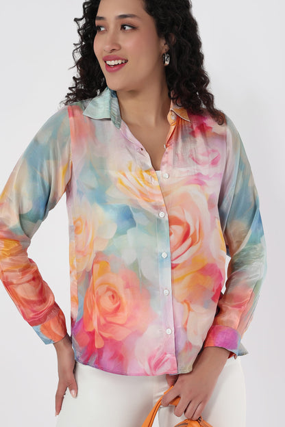 A woman wearing a long-sleeve button-up shirt with a colourful watercolour floral print in pink, orange, yellow, and blue, paired with white pants against a light grey background.