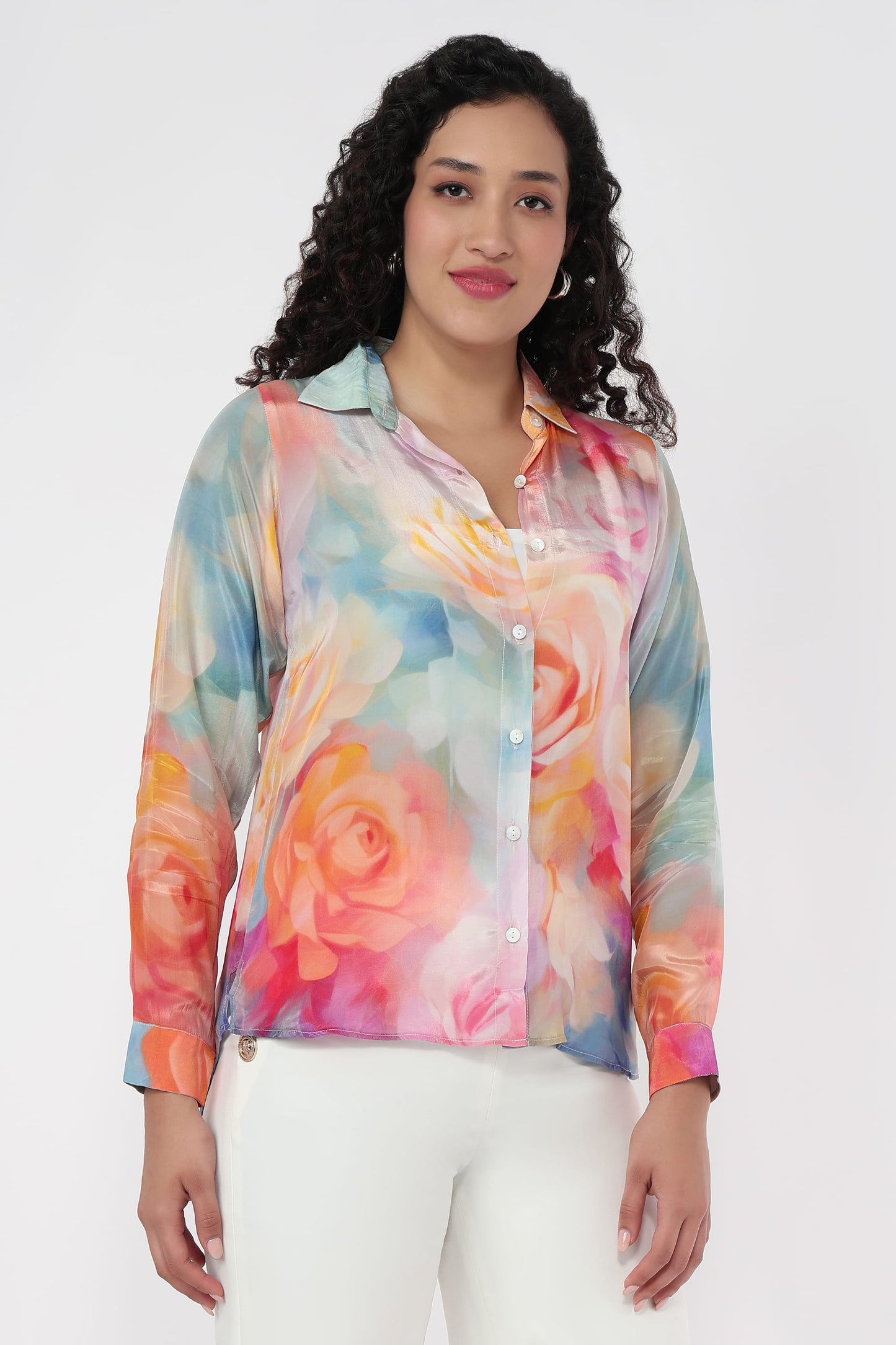 A woman wearing a long-sleeve button-up shirt with a colourful watercolour floral print, paired with white pants, against a plain light grey background.