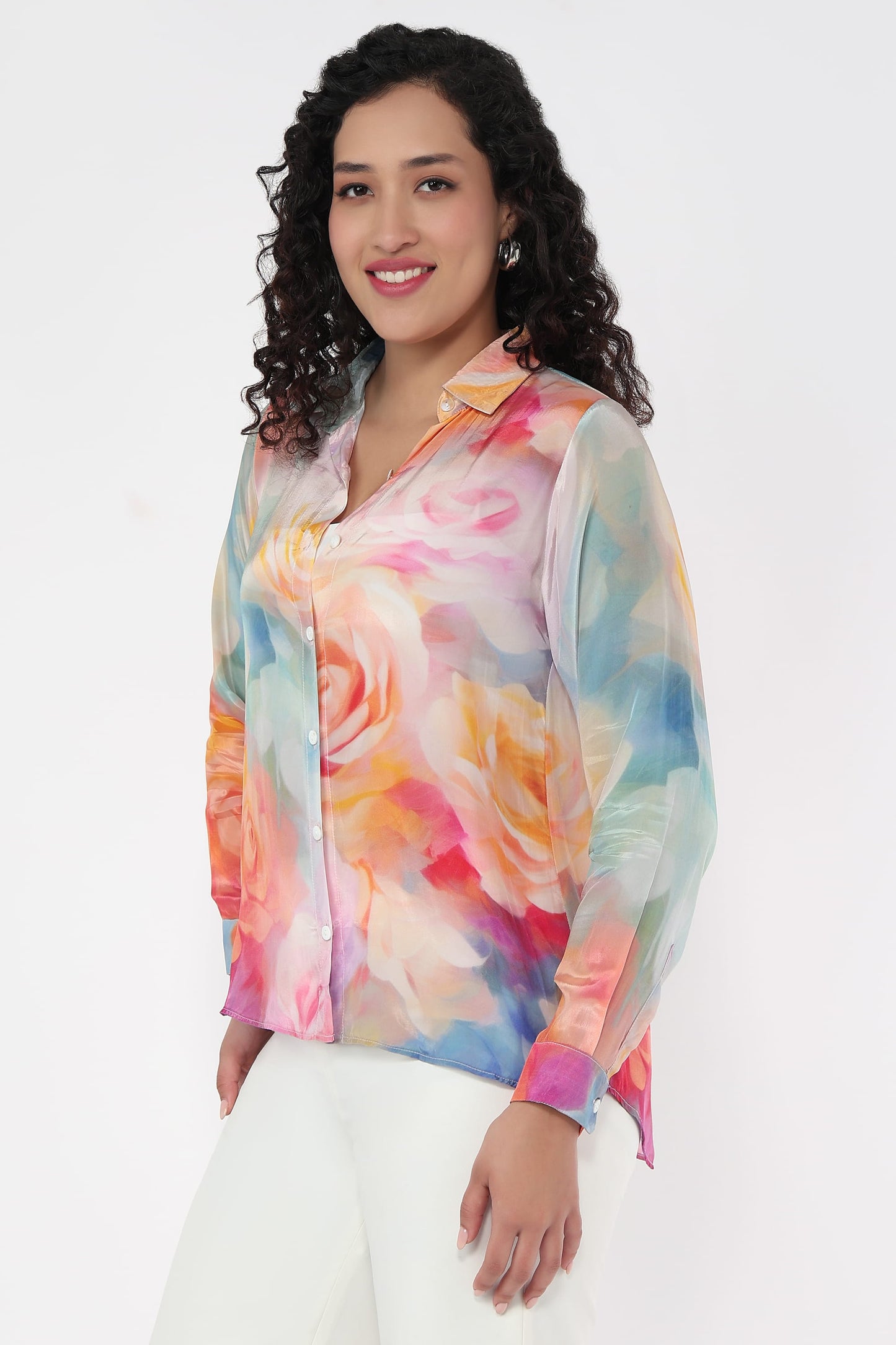 A woman wearing a long-sleeve button-up shirt with a vibrant watercolour floral print in shades of pink, orange, yellow, and blue, paired with white pants, set against a plain white background.