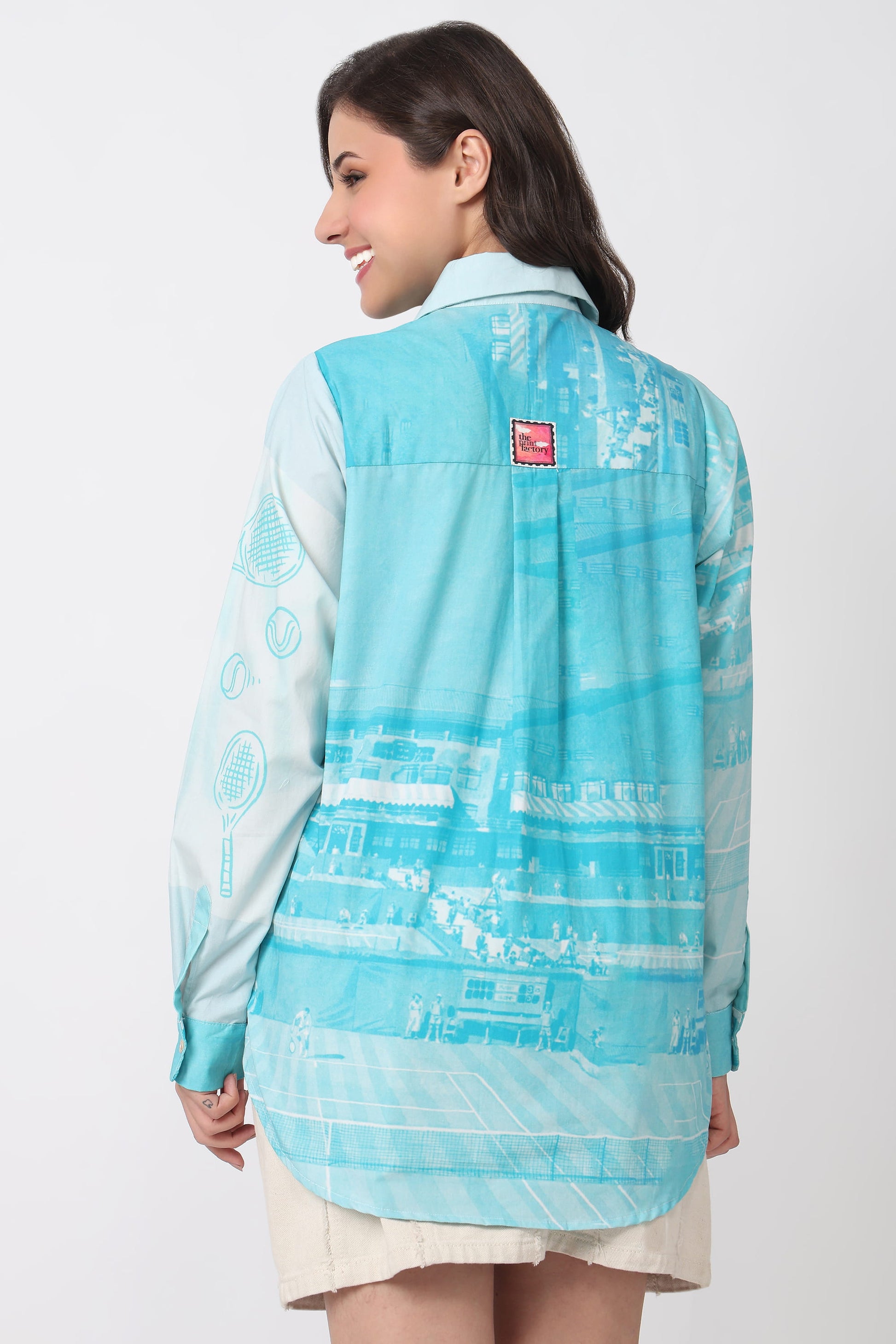A woman shown from the back wearing a light blue and turquoise patterned button-up shirt with a small red square logo patch on the upper back, paired with a light beige skirt, against a plain light gray studio background.
