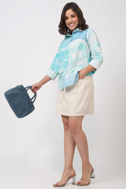 A woman modeling a light blue and white patterned button-up shirt paired with a beige mini skirt, holding a blue handbag against a plain light gray studio background.