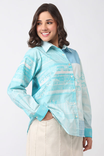 A woman modeling a light blue and white patterned long-sleeve button-up shirt with a solid light blue collar and cuffs, paired with beige trousers, against a plain light gray background.