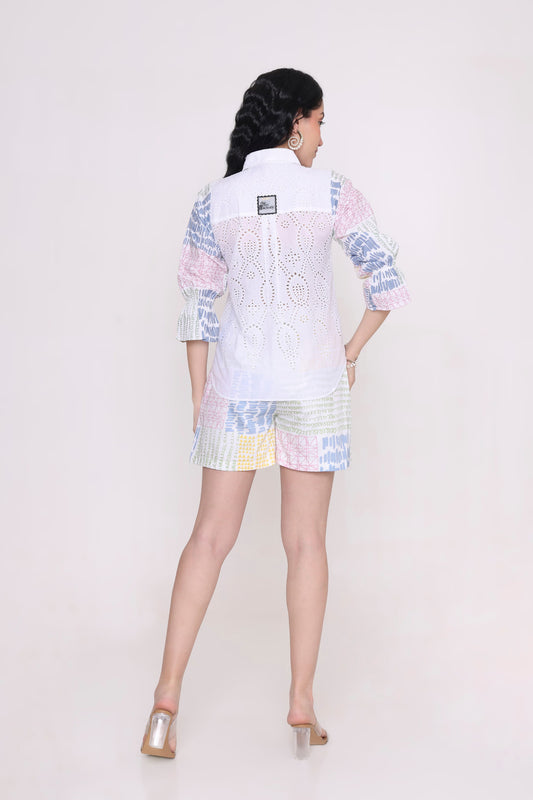 A woman shown from the back wearing a pastel patchwork two-piece set consisting of a white embroidered long-sleeve top and matching patchwork shorts, paired with metallic heeled sandals, against a plain light background.