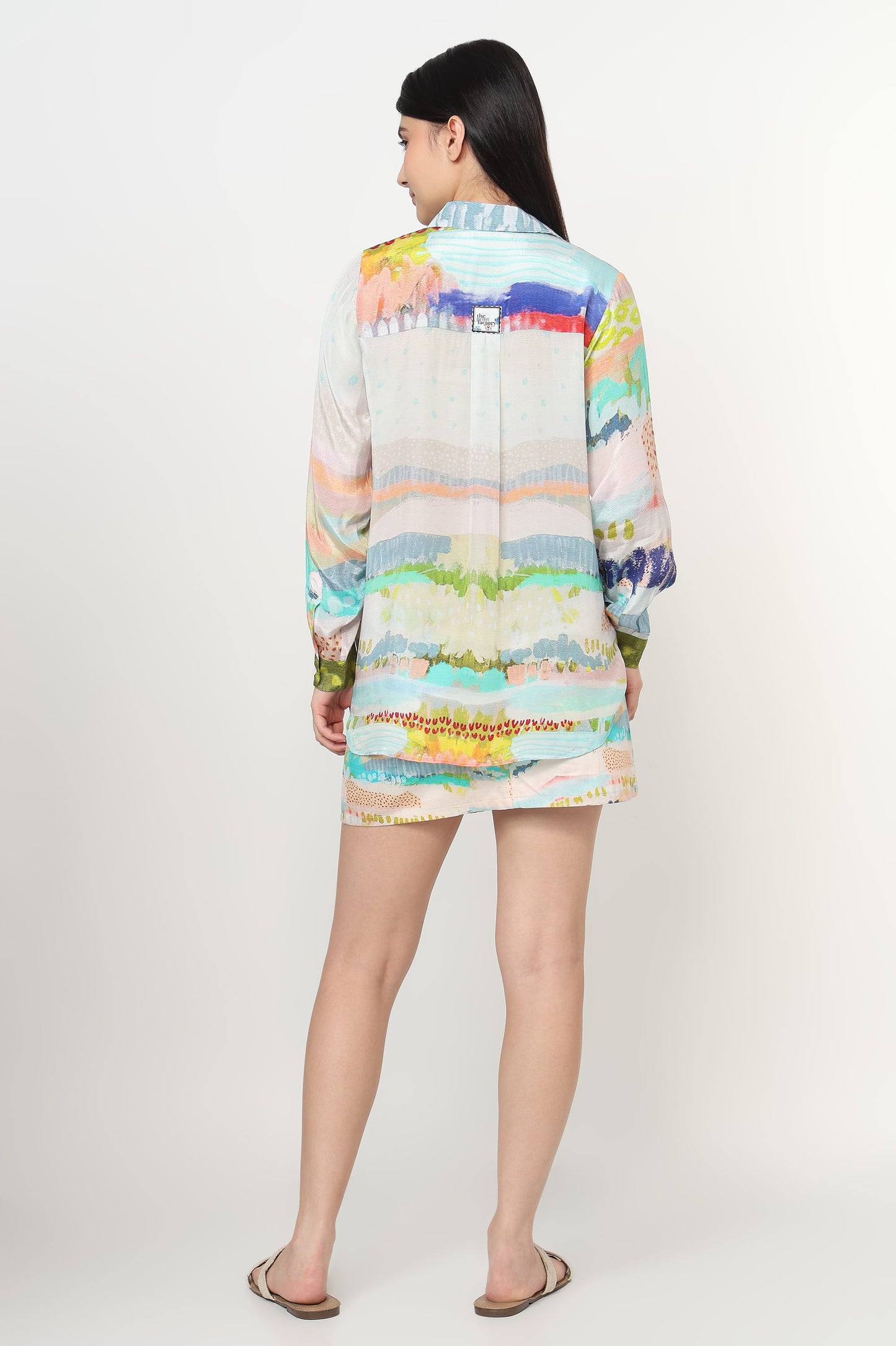 A woman shown from the back wearing a long-sleeve,co-ord set in pastel shades of blue, green, yellow, orange, and pink on a white base, photographed against a plain light gray studio background.