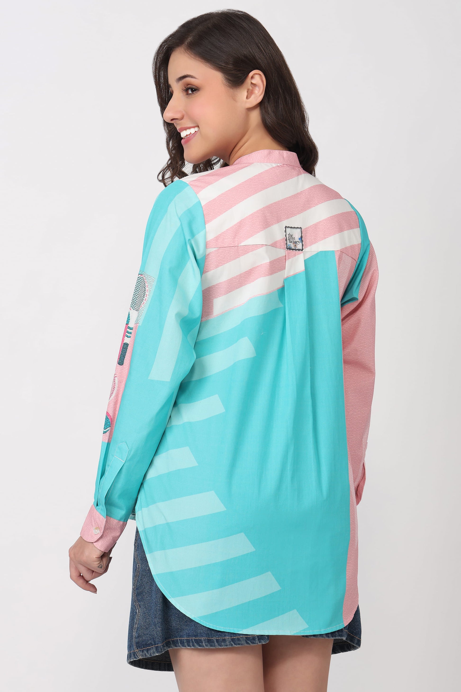 A woman modeling a long-sleeve colorblock shirt with a turquoise front panel, pink and white striped yoke, and pink sleeves, shown from a three-quarter back view against a light gray studio background.