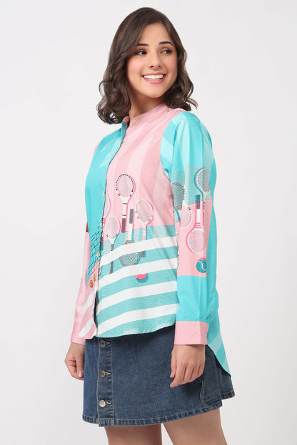 A woman modeling a long-sleeve collared top with a pink and turquoise color-blocked design, white stripes, and tennis racket graphics, paired with a denim skirt against a light gray background.