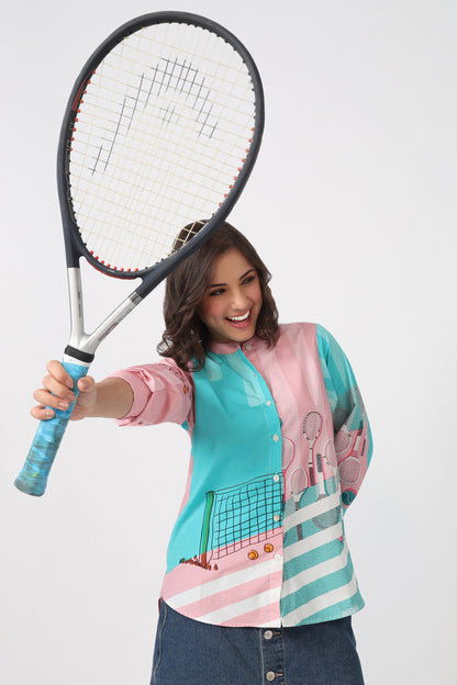 A woman holding a tennis racket, wearing a colorful long-sleeve button-up shirt with a tennis-themed print in pink, teal, and white, and a denim skirt, against a light gray background.