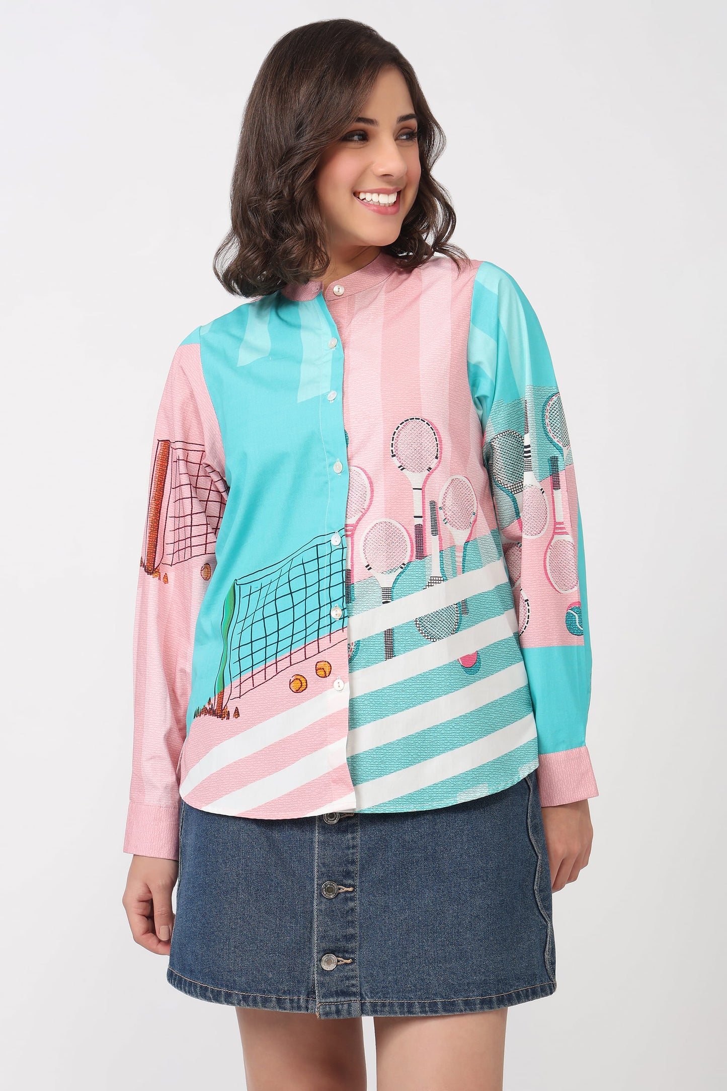 A woman modeling a long-sleeve button-up shirt with a colorful tennis-themed print in pink, turquoise, and white, paired with a denim mini skirt against a light gray studio background.