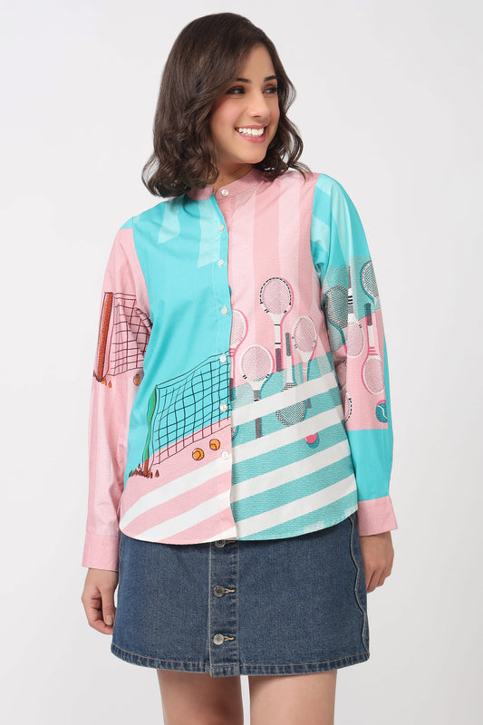 A woman modeling a long-sleeve button-up shirt with a colorful tennis-themed print in pink, turquoise, and white, paired with a denim mini skirt against a light gray studio background.