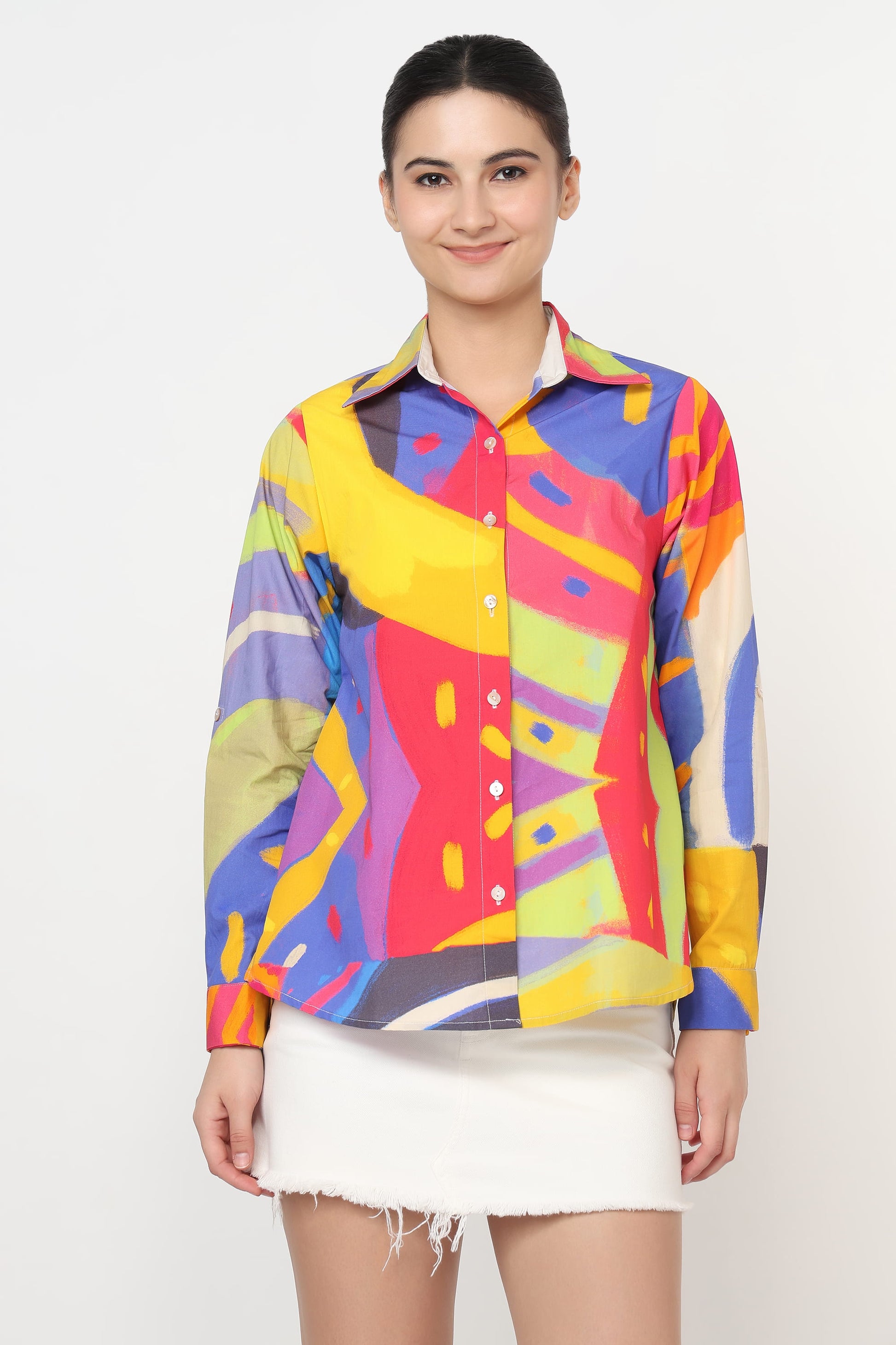 A woman modeling a long-sleeve button-up shirt with a vibrant, multicolored abstract geometric print, paired with a white skirt, against a plain light gray background.