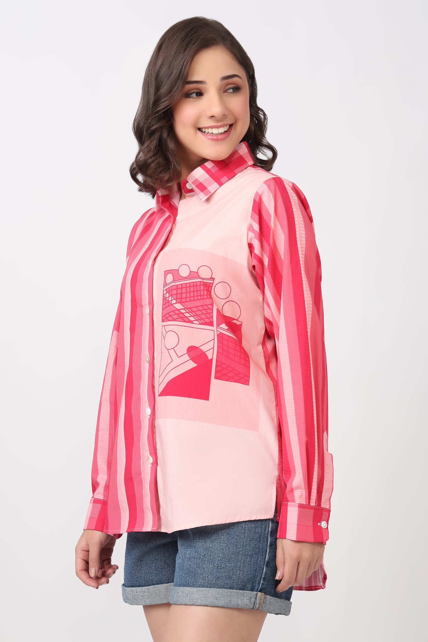 A woman modeling a pink and red striped button-up shirt with a pink graphic print on the front, paired with denim shorts, against a light gray studio background.
