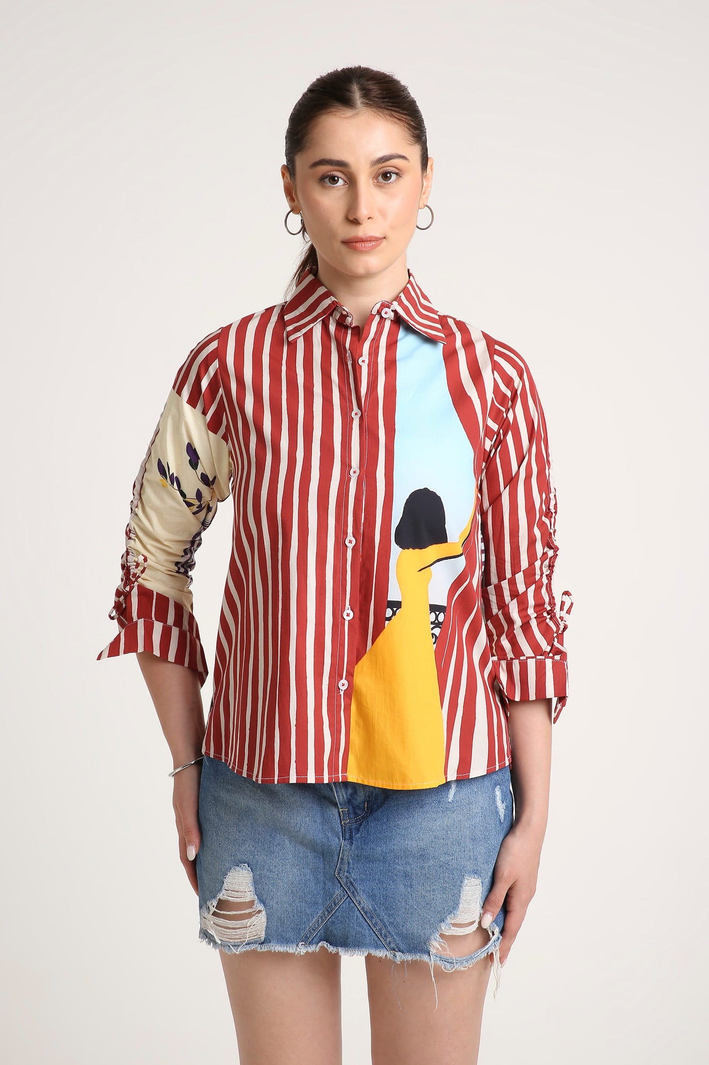 A woman wearing a long-sleeve button-up shirt with red and white vertical stripes, a beige patterned right sleeve, and a colourful front graphic featuring a yellow illustration and a light blue background, paired with a denim skirt against a light grey studio backdrop.