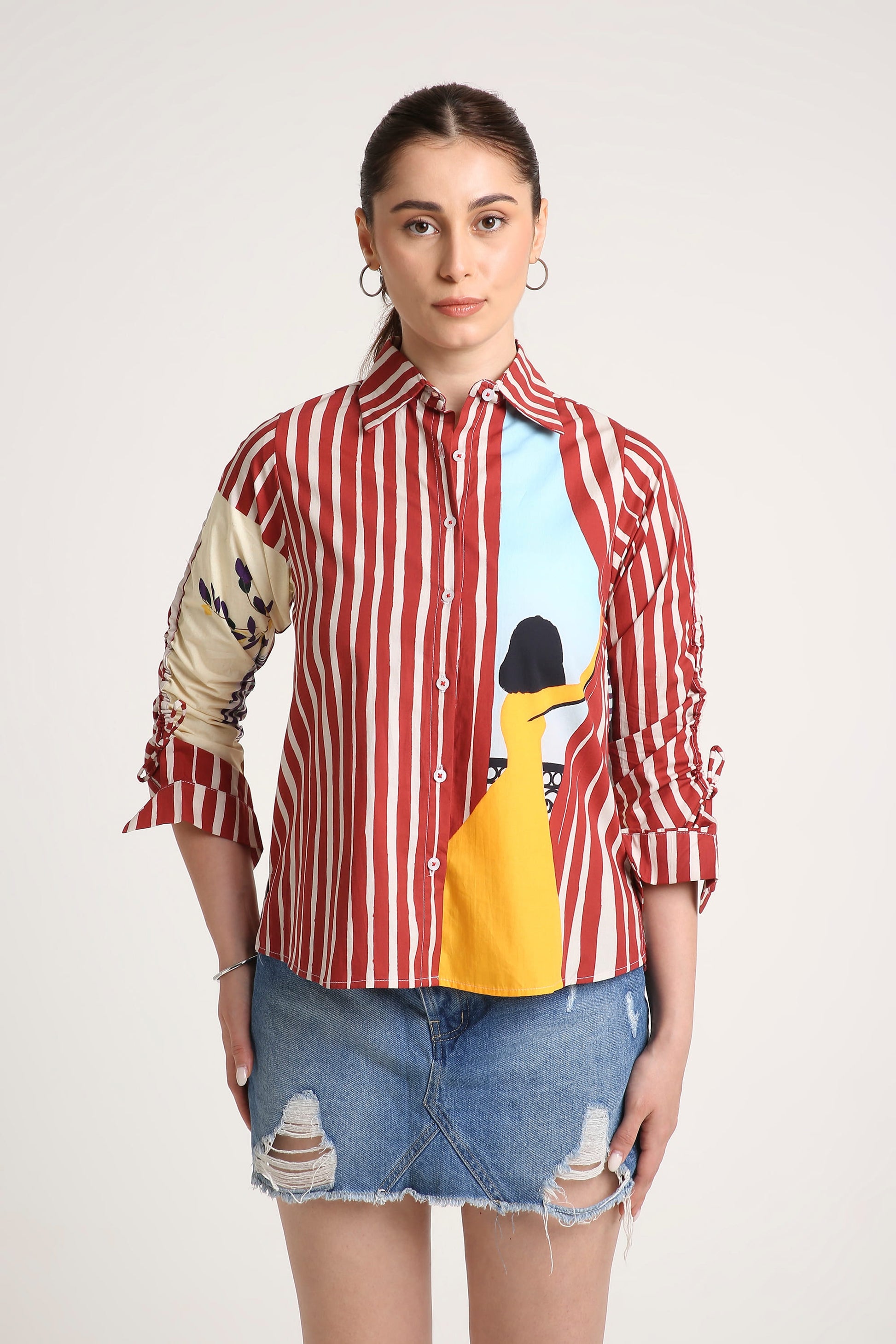 A woman wearing a long-sleeve button-up shirt with red and white vertical stripes, a beige patterned right sleeve, and a colourful front graphic featuring a yellow illustration and a light blue background, paired with a denim skirt against a light grey studio backdrop.