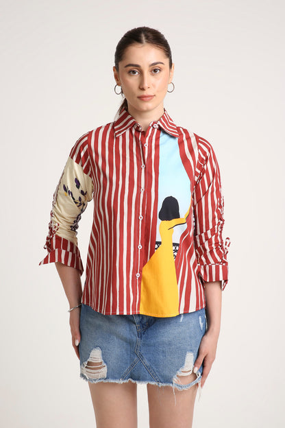 A woman wearing a long-sleeve button-up shirt with red and white vertical stripes, a beige patterned right sleeve, and a colourful front graphic featuring a yellow illustration and a light blue background, paired with a denim skirt against a light grey studio backdrop.