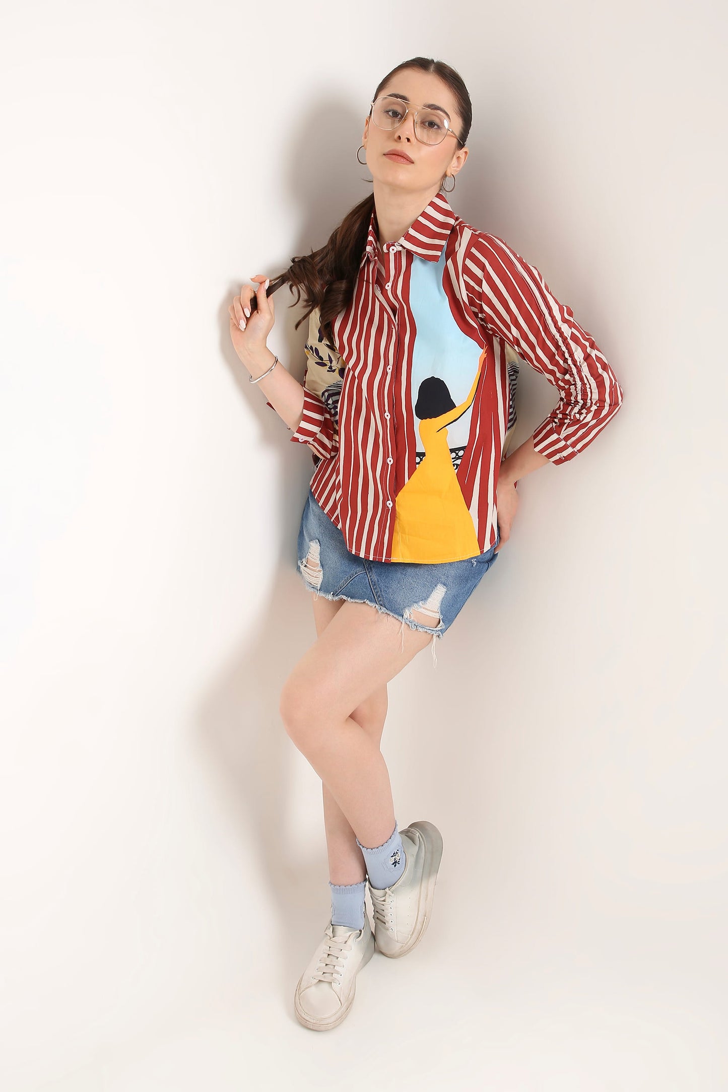 A woman wearing a long-sleeve button-up shirt with red and white vertical stripes, a beige patterned right sleeve, and a colourful front graphic featuring a yellow illustration and a light blue background, paired with a denim skirt against a light grey studio backdrop.