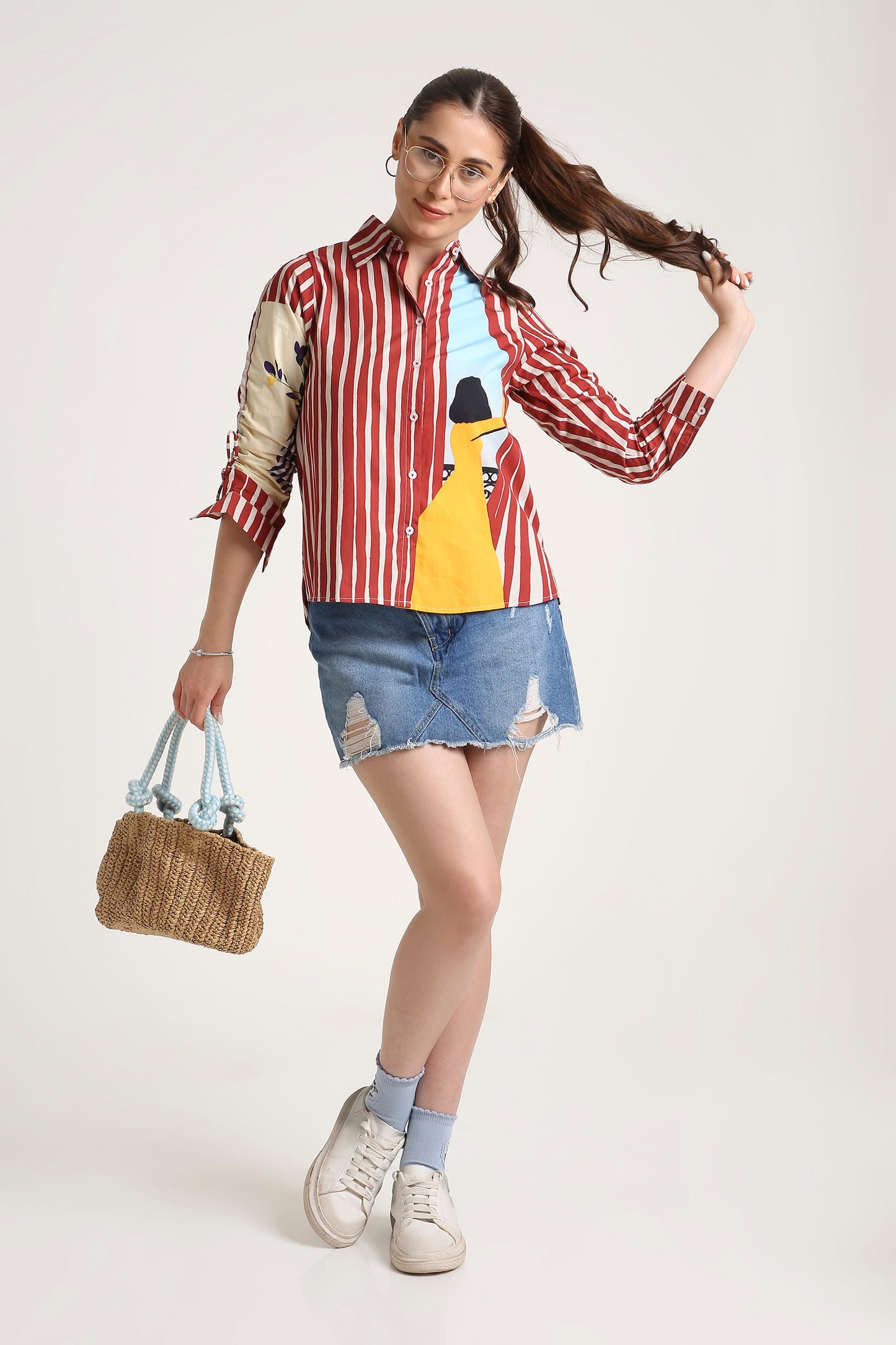 A woman modeling a red and white vertically striped button-up shirt with a graphic print on the front, paired with a denim mini skirt, white sneakers, and holding a woven straw handbag against a plain light background.