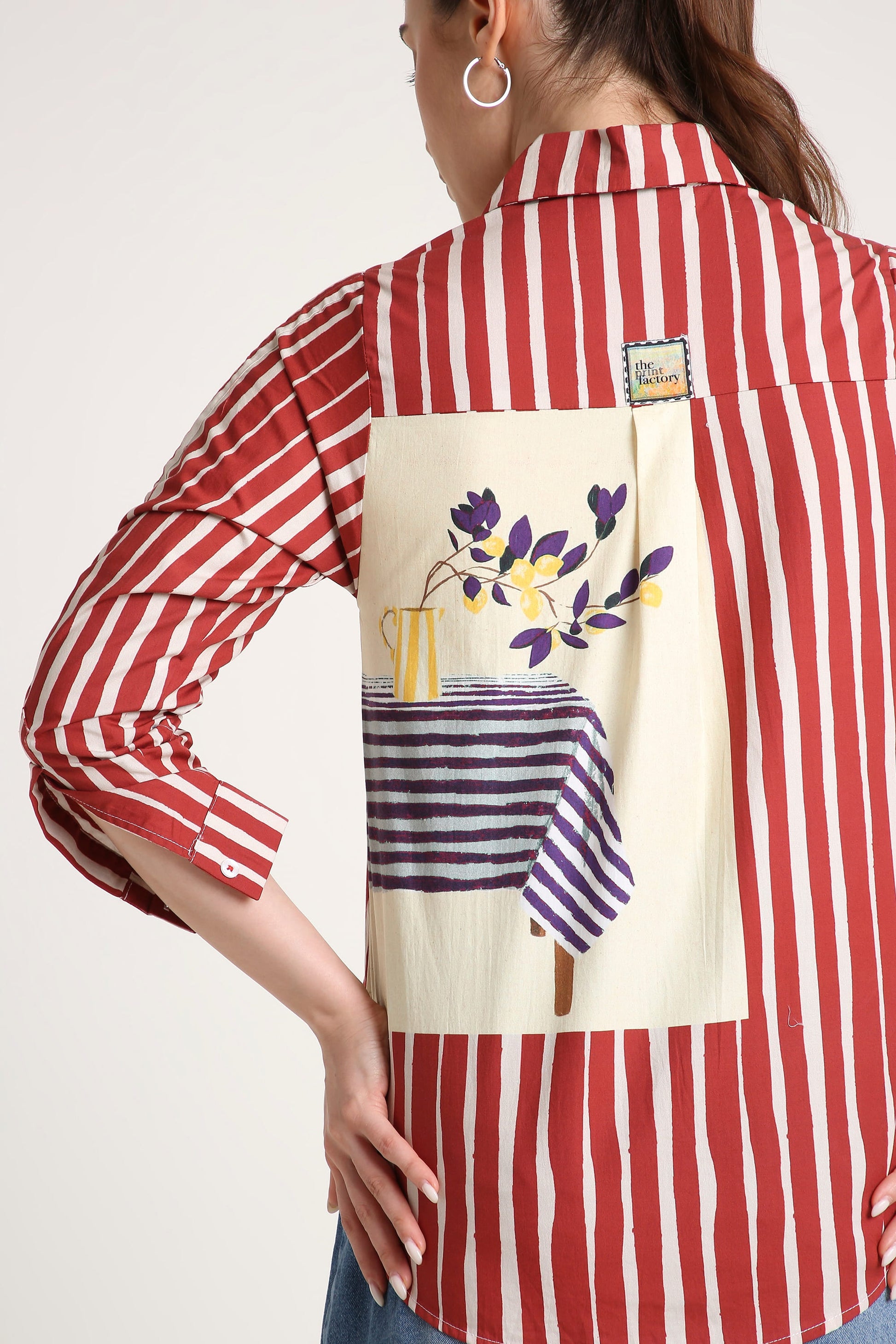 A woman shown from the back wearing a red and white vertically striped button-up shirt with a large cream patch on the back featuring a floral still life design, photographed against a light gray studio background.