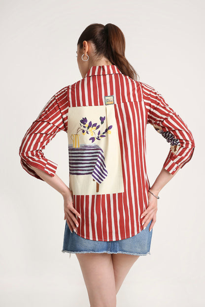 A woman shown from the back wearing a red and white vertically striped button-up shirt with a large cream-colored graphic print on the back, paired with a denim skirt, against a plain light background.