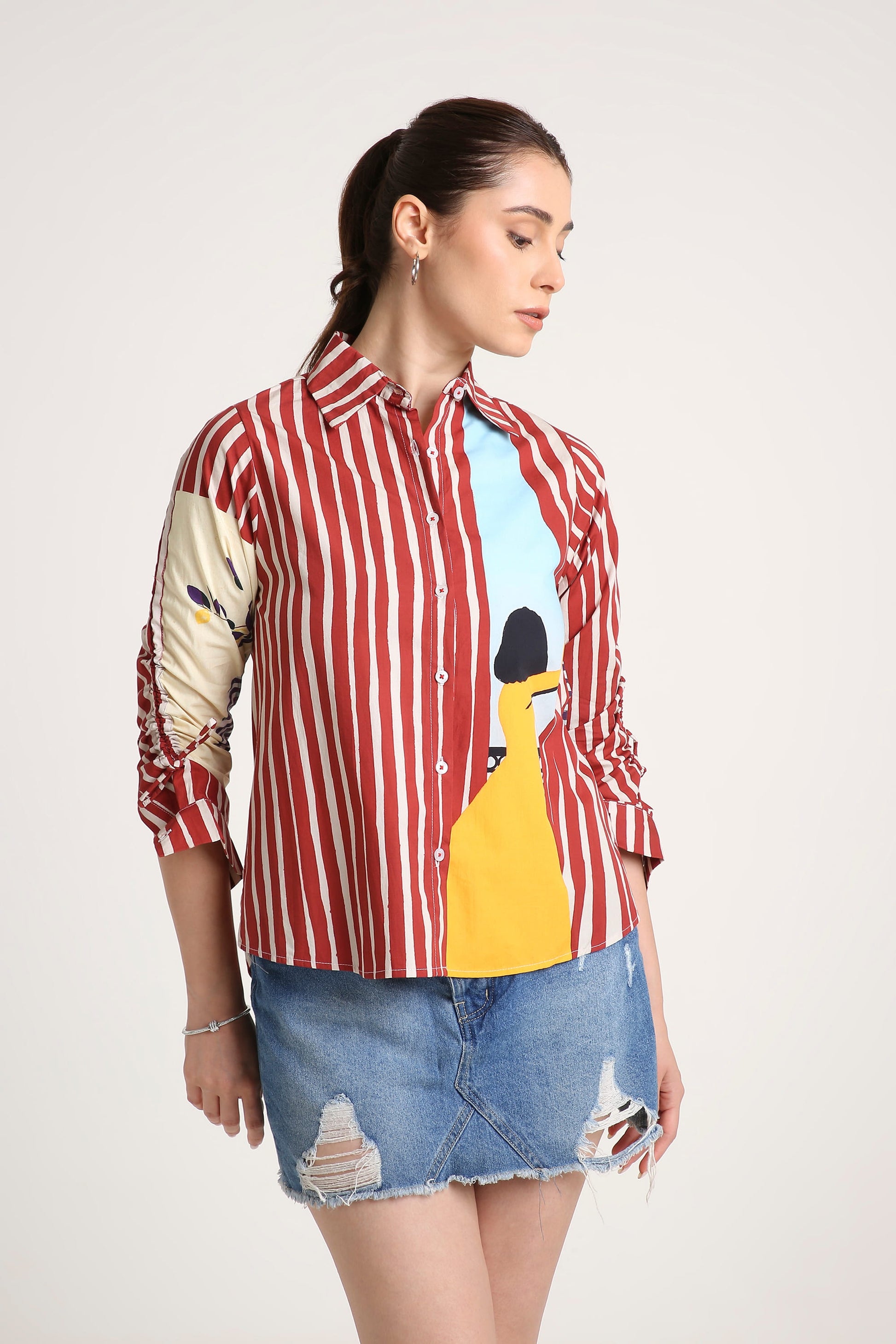 A woman modeling a red and white vertically striped button-up shirt with a graphic print on the front and sleeves, paired with a distressed denim mini skirt, against a light gray studio background.