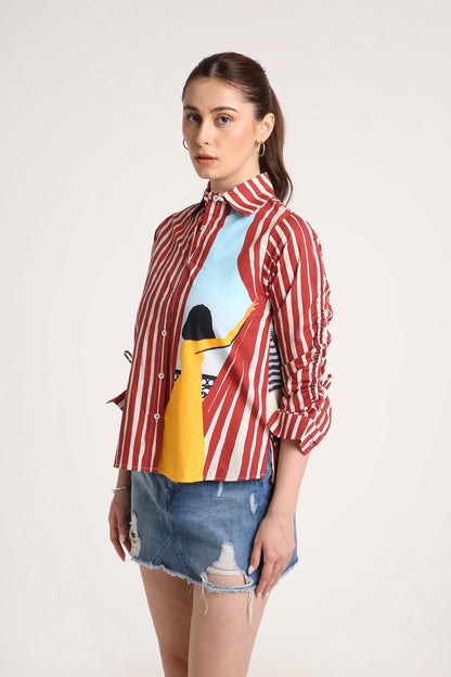 A woman wearing a long-sleeve button-up shirt with red and white vertical stripes, a beige patterned right sleeve, and a colourful front graphic featuring a yellow illustration and a light blue background, paired with a denim skirt against a light grey studio backdrop.