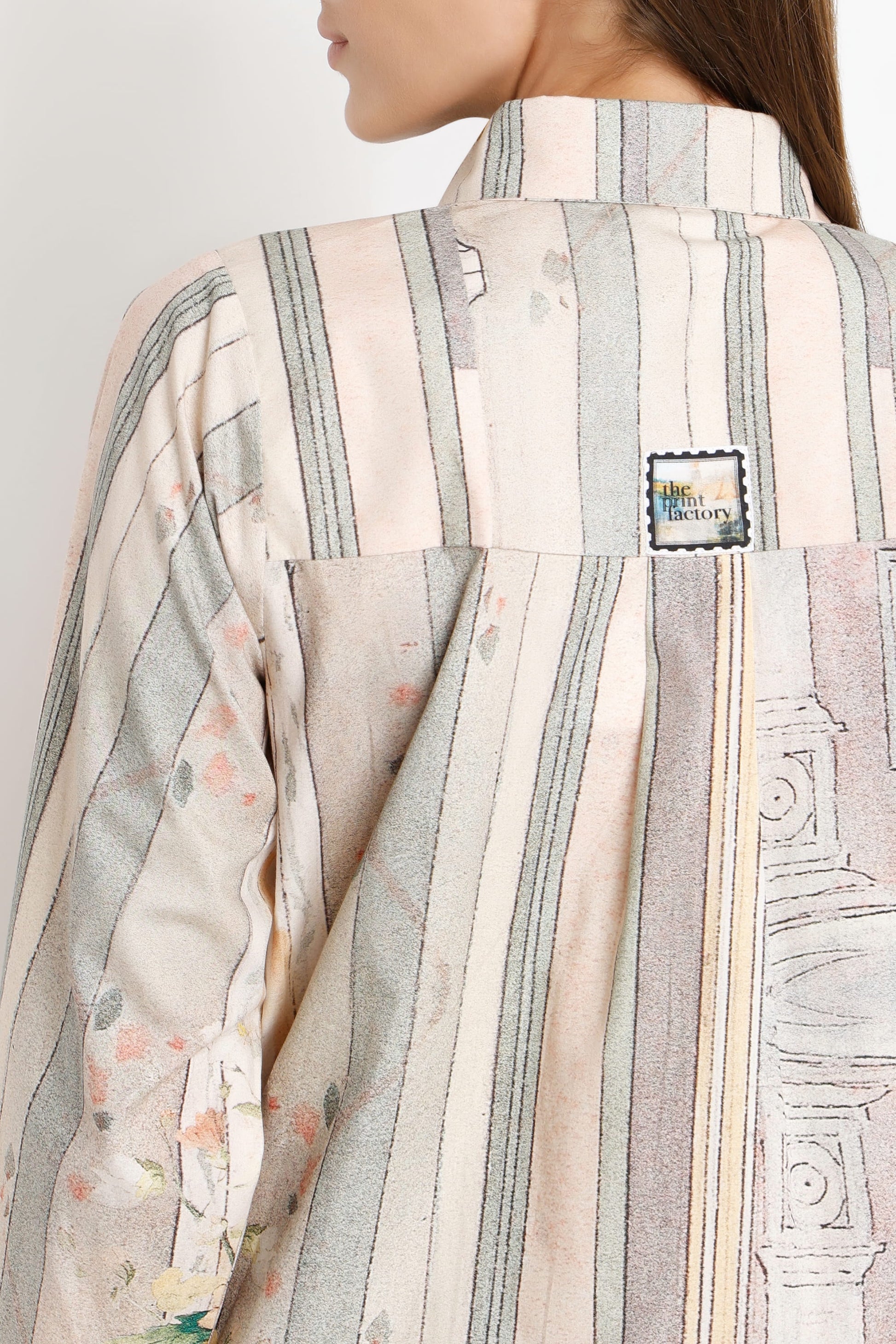 Close-up of a model wearing a striped shirt with vertical bands of beige, gray, and soft pink, featuring a small square brand patch on the upper back.