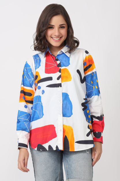 A woman modeling a long-sleeve button-up shirt with a bold abstract geometric print in blue, red, orange, yellow, and black on a white base, paired with light blue jeans against a light gray studio background.