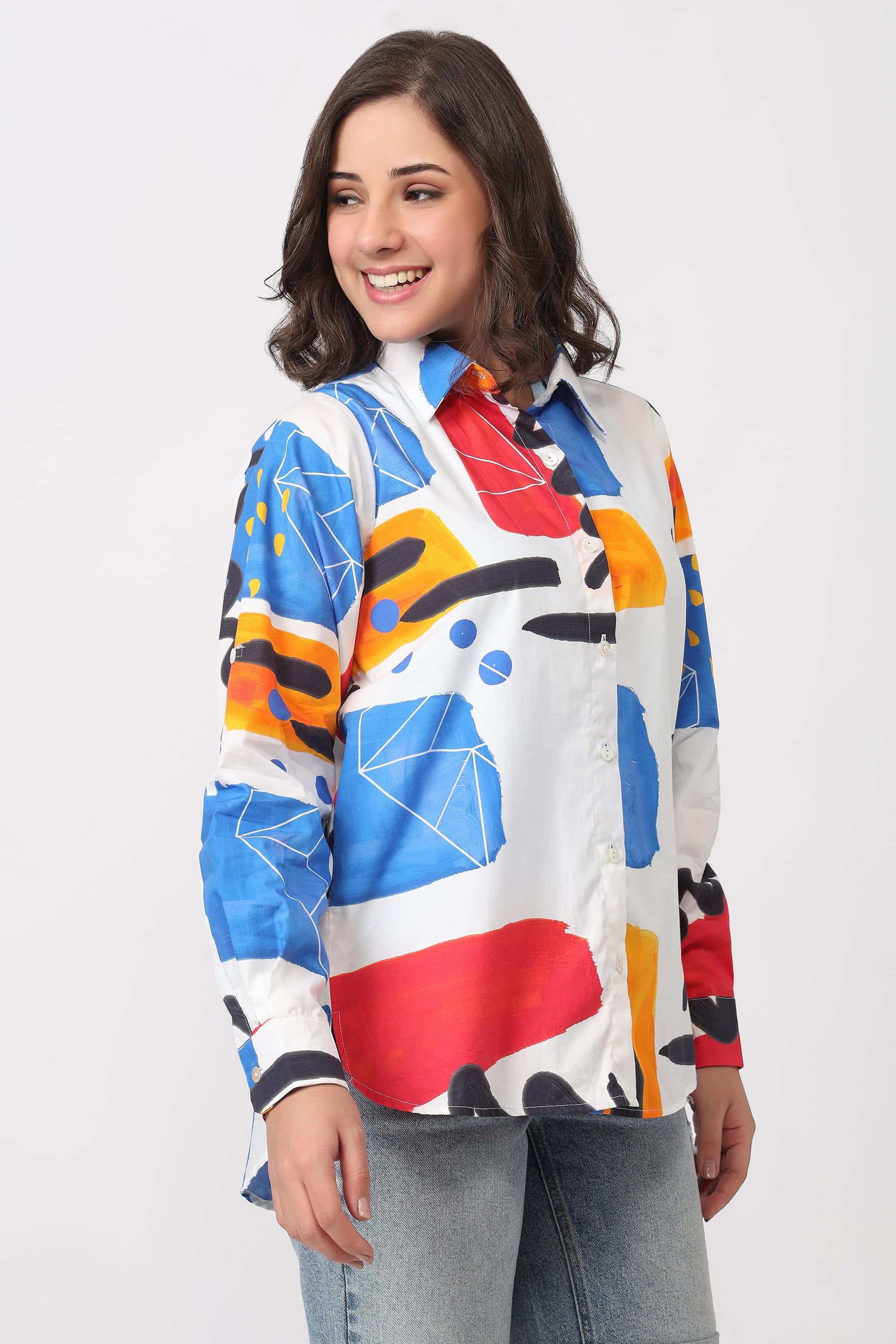 A woman wearing a long-sleeve collared button-up shirt with a bold abstract geometric print in blue, red, orange, yellow, black, and white, paired with light-wash jeans against a light gray studio background.