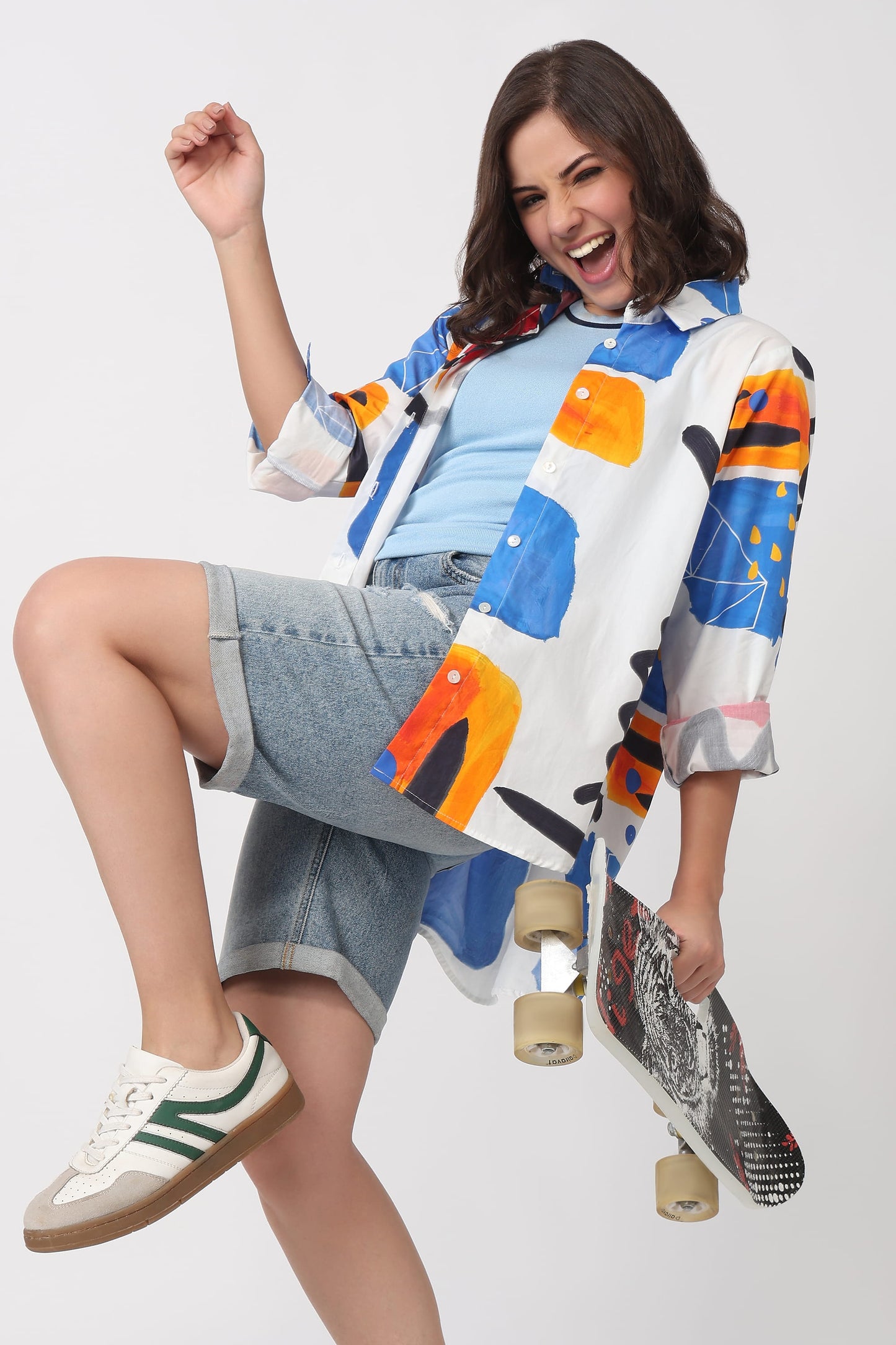 A woman in a white button-up shirt with a colorful abstract geometric print, a light blue t-shirt, denim shorts, and white sneakers with green stripes, holding a skateboard against a light gray studio background.