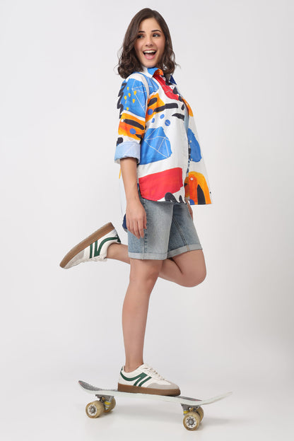 A woman posing on a skateboard against a light gray studio background, wearing a colorful abstract print short-sleeve shirt, denim shorts, and white sneakers with green stripes.
