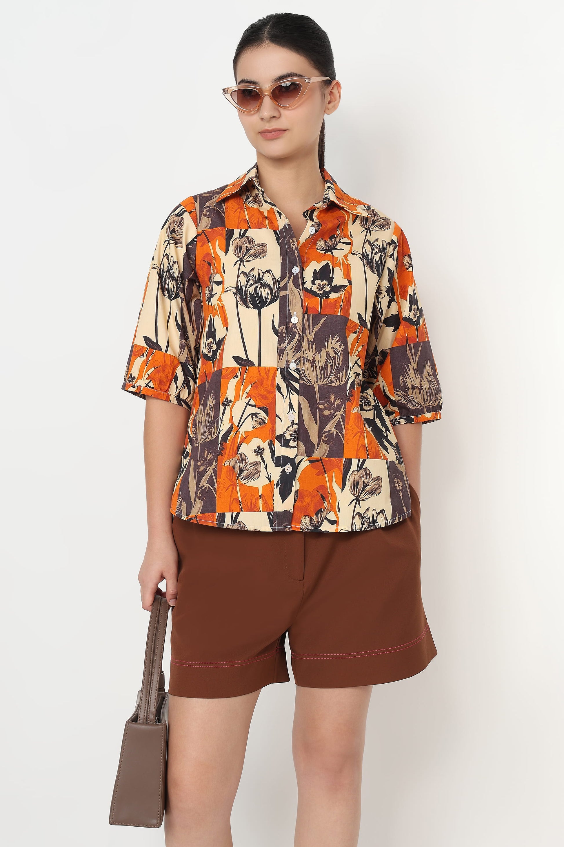 Woman wearing a patterned shirt and brown shorts with sunglasses on a white background