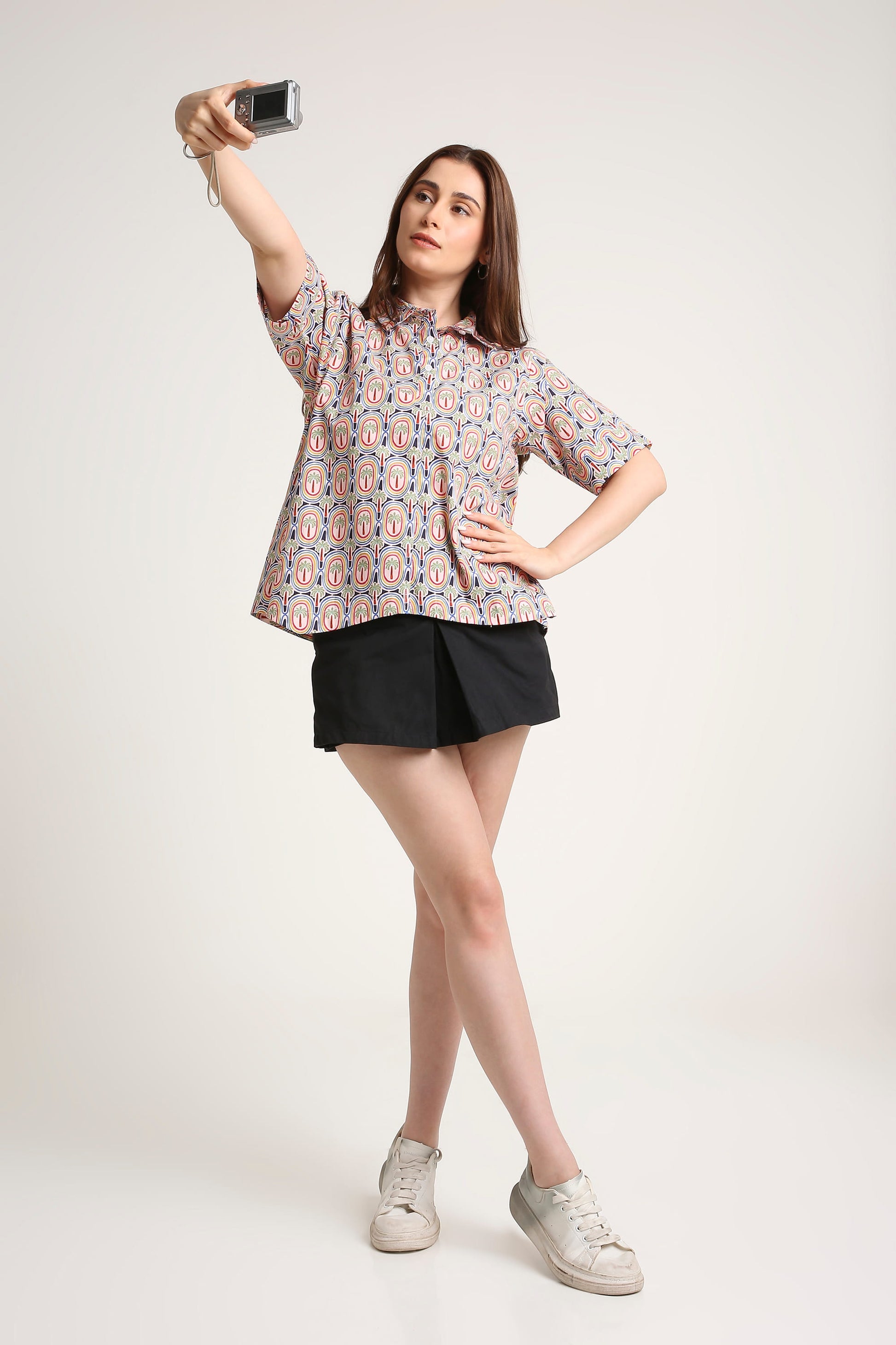 A woman posing for a mirror selfie in a studio setting, wearing a short-sleeve button-up shirt with a pink, blue, and white pattern, black shorts, and white sneakers.