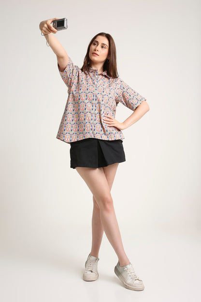 A woman posing for a mirror selfie in a studio setting, wearing a short-sleeve button-up shirt with a pink, blue, and white pattern, black shorts, and white sneakers.