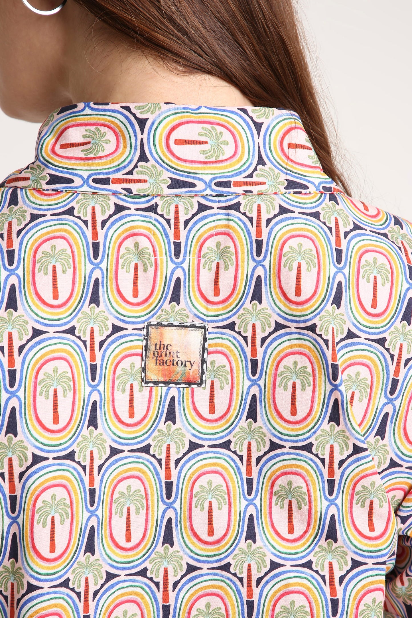 Close-up of a person wearing a collared button-up shirt with a colorful retro pattern of palm trees inside rainbow arches on a navy background. A square brand patch reading 'the print factory' is visible on the back.