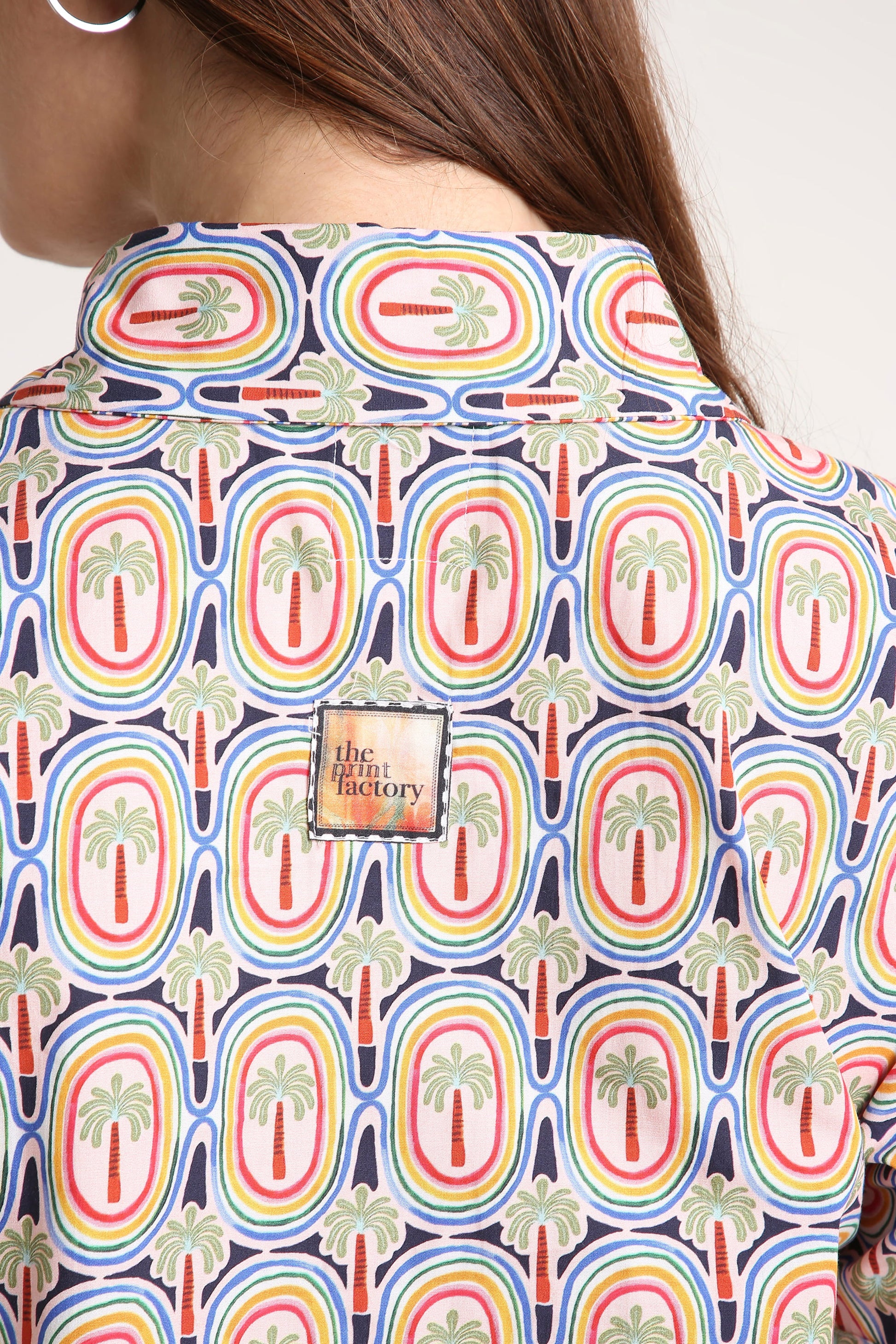 Close-up of a person wearing a collared button-up shirt with a colorful retro pattern of palm trees inside rainbow arches on a navy background. A square brand patch reading 'the print factory' is visible on the back.