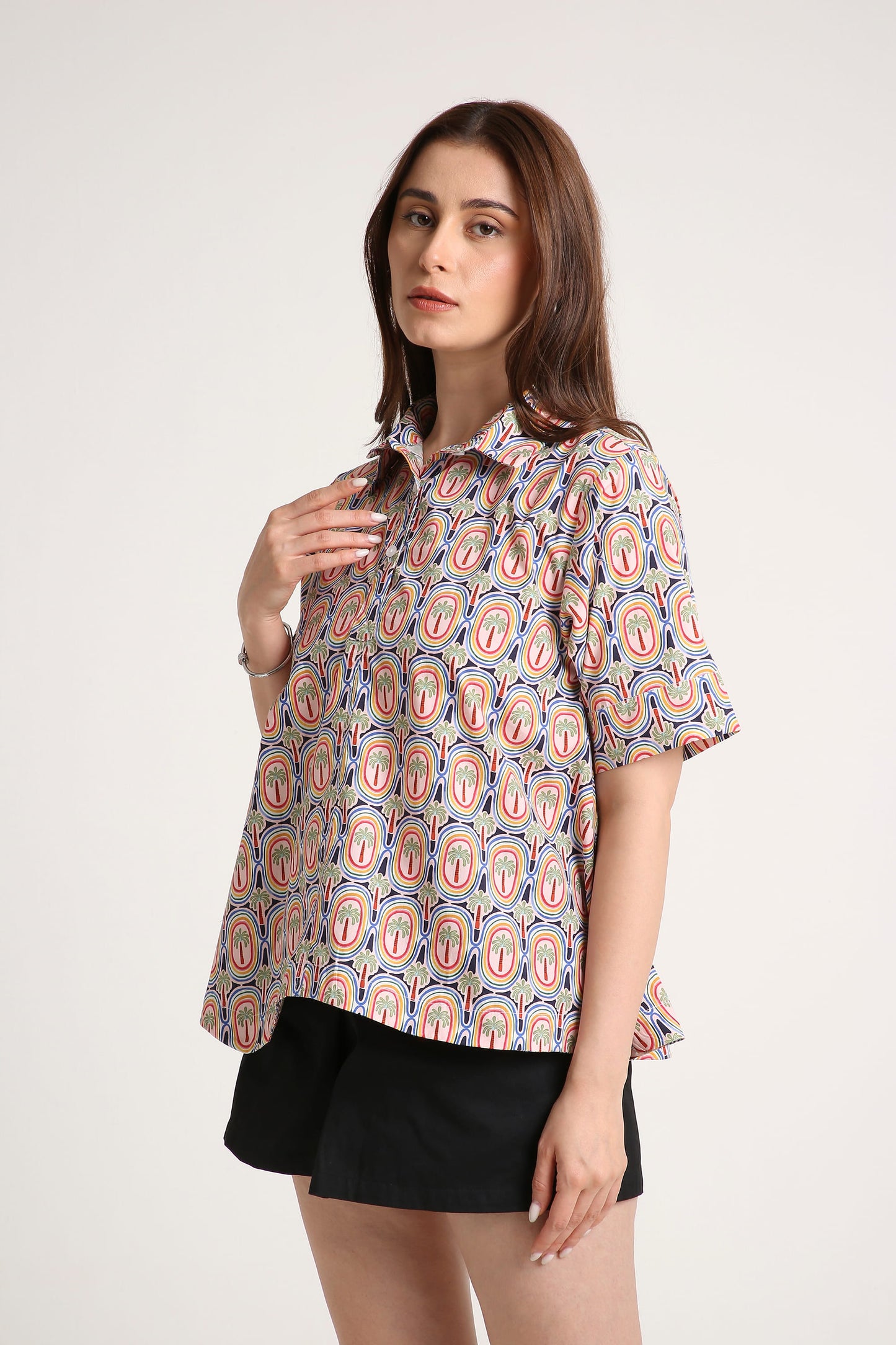 A woman wearing a short-sleeve button-up shirt with a colourful paisley pattern in red, orange, and blue on a light background, paired with black shorts, against a plain light grey studio background.
