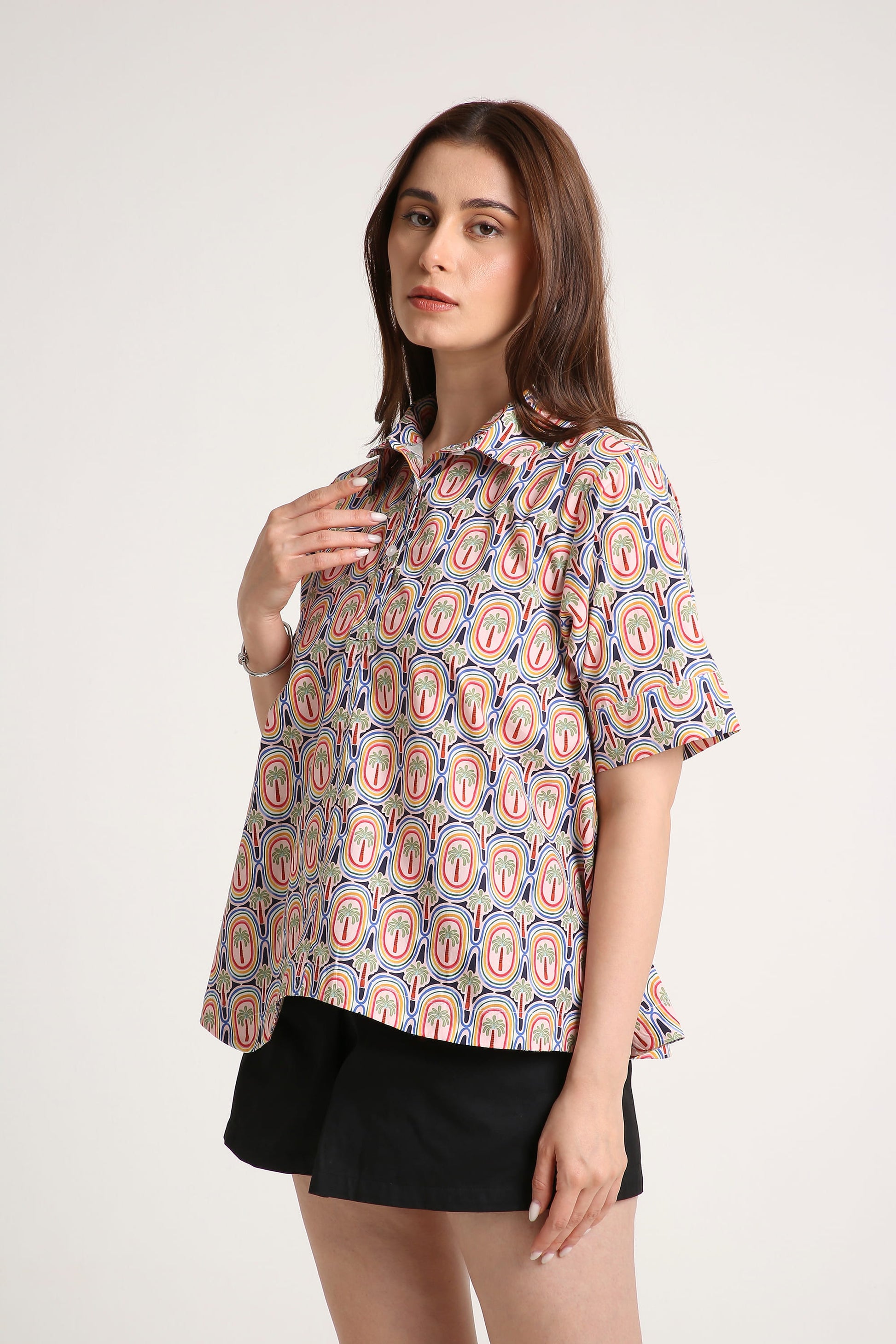 A woman wearing a short-sleeve button-up shirt with a colourful paisley pattern in red, orange, and blue on a light background, paired with black shorts, against a plain light grey studio background.