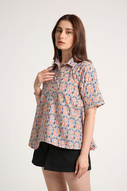 A woman wearing a short-sleeve button-up shirt with a colourful paisley pattern in red, orange, and blue on a light background, paired with black shorts, against a plain light grey studio background.