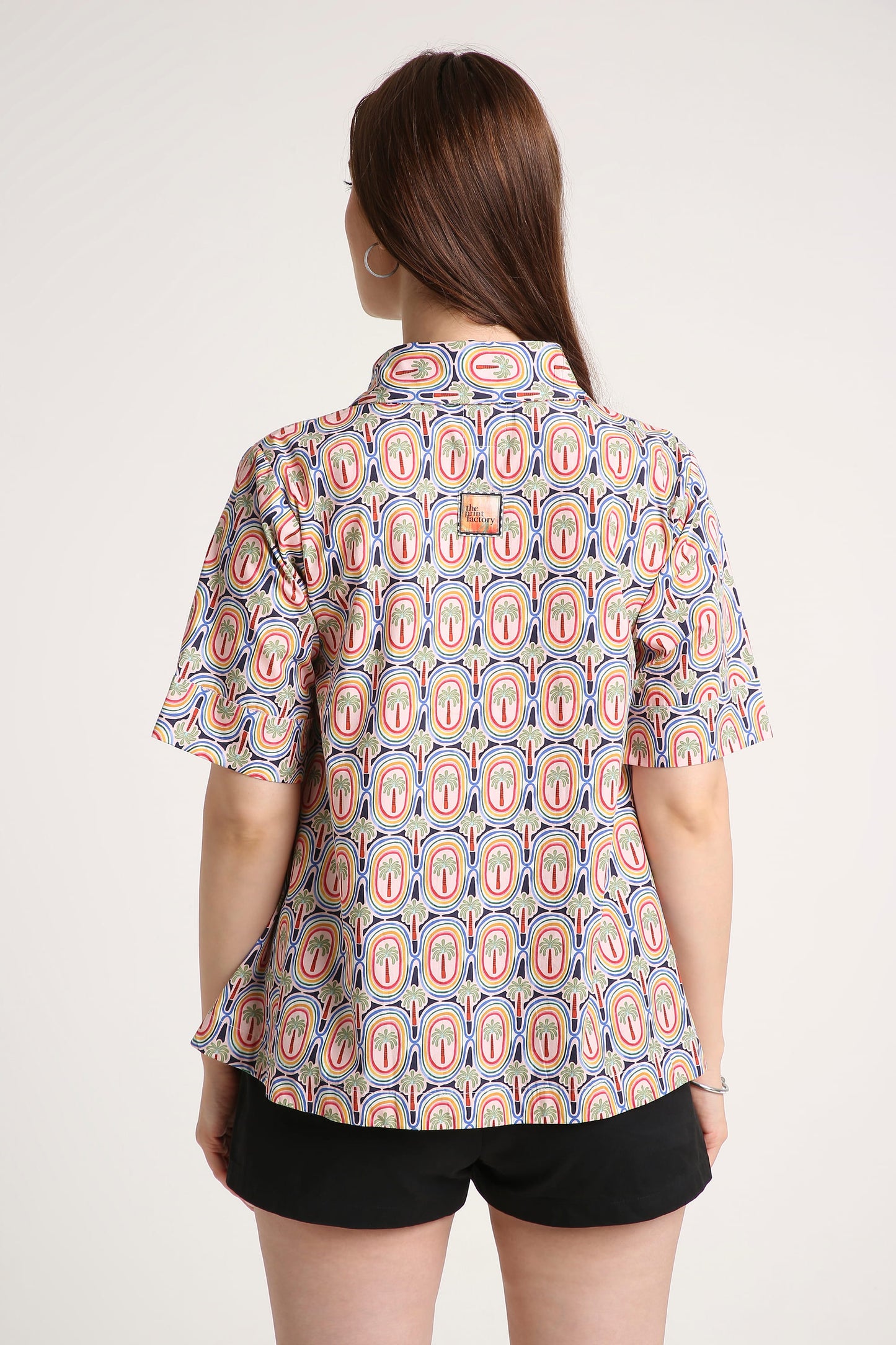A woman viewed from the back wearing a short-sleeve button-up shirt with a repeating paisley-like oval pattern in pink, orange, and blue on a light base, paired with black shorts against a plain light background.