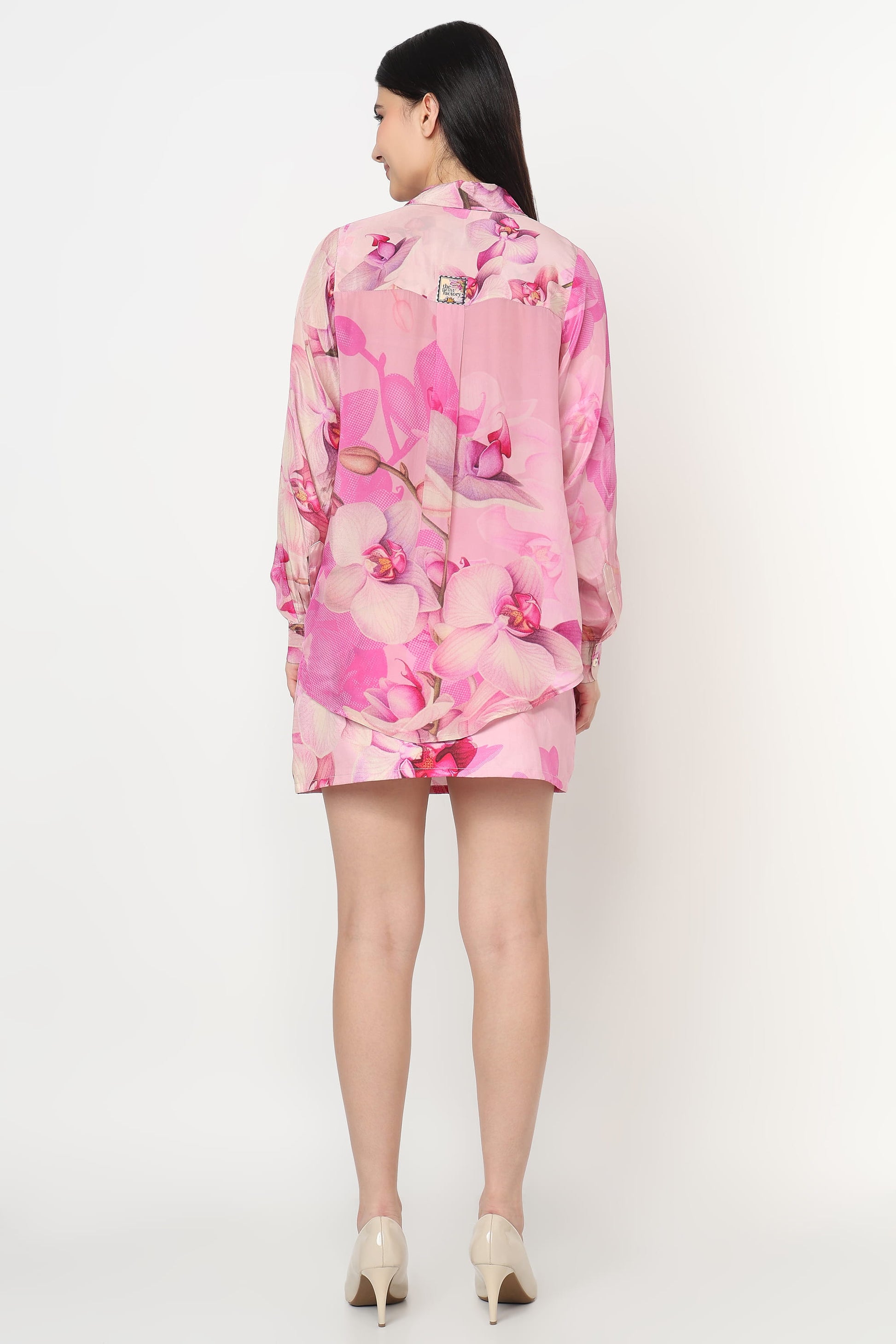 A woman viewed from the back wearing a pink floral long-sleeve shirt dress with a small square patch on the upper back, paired with beige high heels, against a plain white studio background.