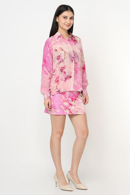 A woman modeling a pink floral two-piece set consisting of a long-sleeve button-up shirt and a matching mini skirt, paired with beige high heels, against a plain white studio background.